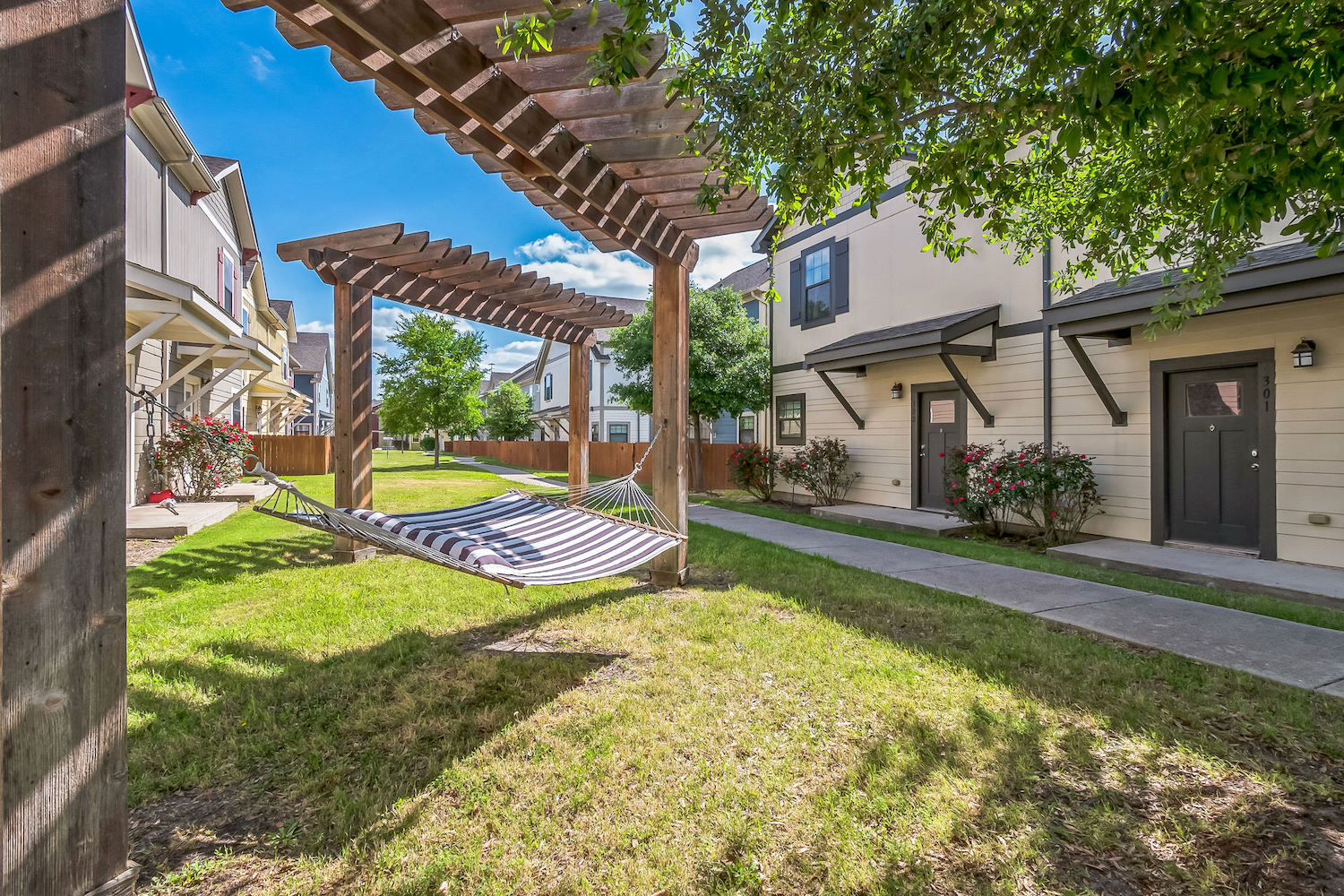 A striped hammock hangs under a wooden pergola on a grassy lawn between two rows of modern apartments in College Station, with trees and bushes adding greenery to the scene.