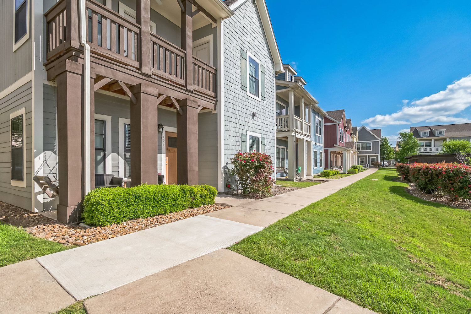 Row of modern townhouses with balconies and covered porches at The Heights College Station, bordered by neatly trimmed bushes and a green lawn, on a sunny day with a blue sky and scattered clouds.