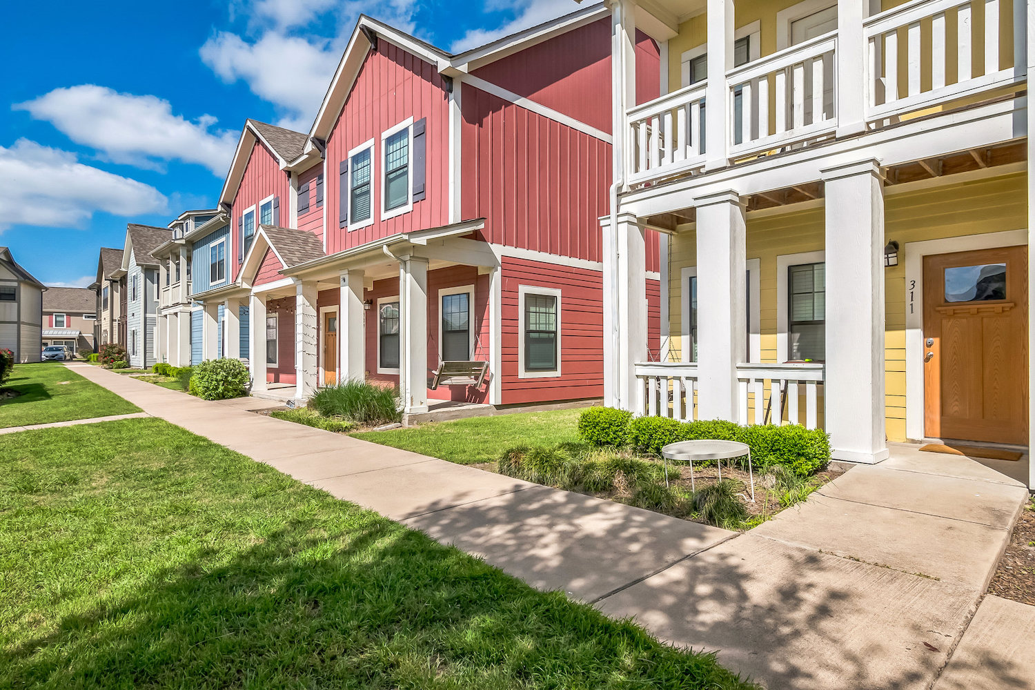 Colorful row of modern townhouses with porches and well-kept lawns, these apartments in College Station line a sidewalk under a bright blue sky with scattered clouds.