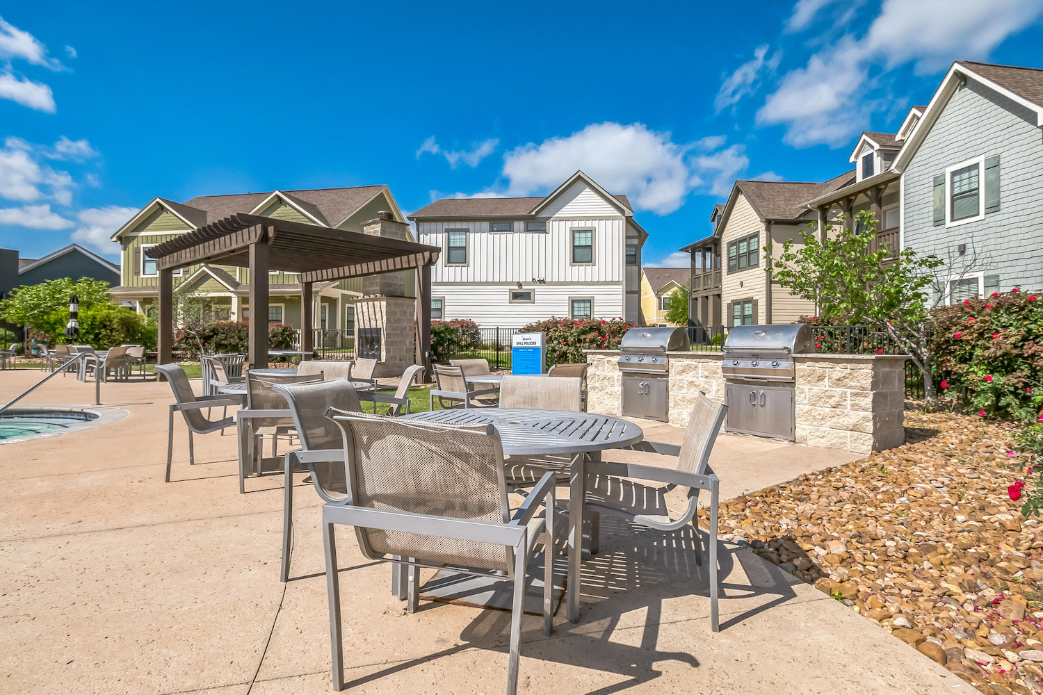 Outdoor patio area with metal tables and chairs, a covered pergola, barbecue grills, and landscaping, set among modern suburban houses under a blue sky—perfect for relaxing at tamu off campus housing.