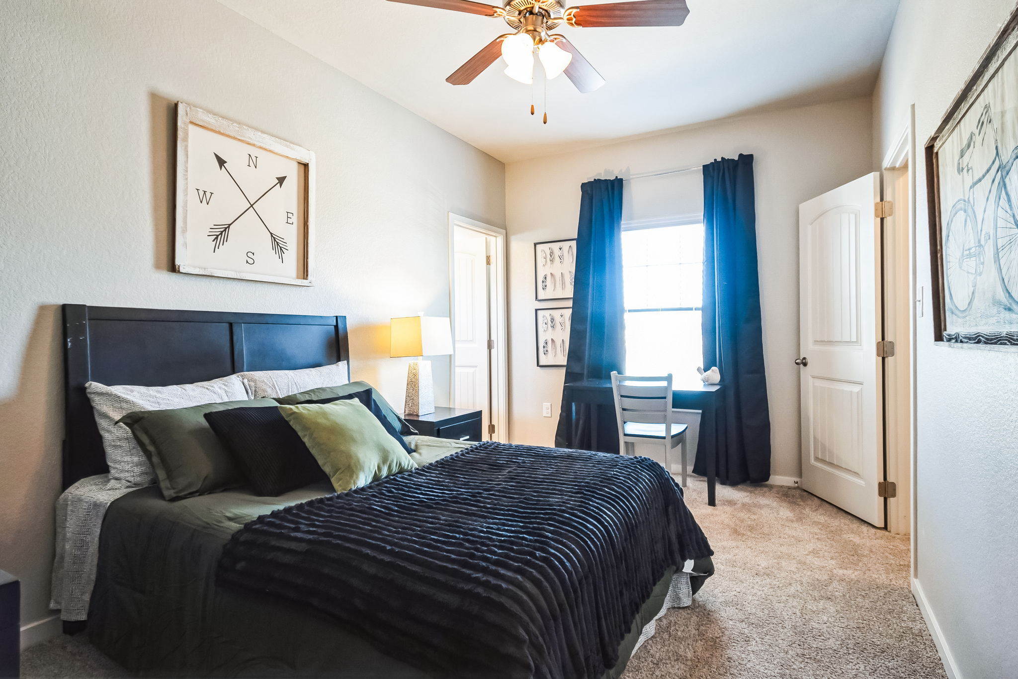 A cozy bedroom in tamu off campus housing features a black bed with green pillows and a black blanket. A ceiling fan, lamp, and wall art enhance the space. There’s a small desk by the window with dark curtains, and a door is partially open.