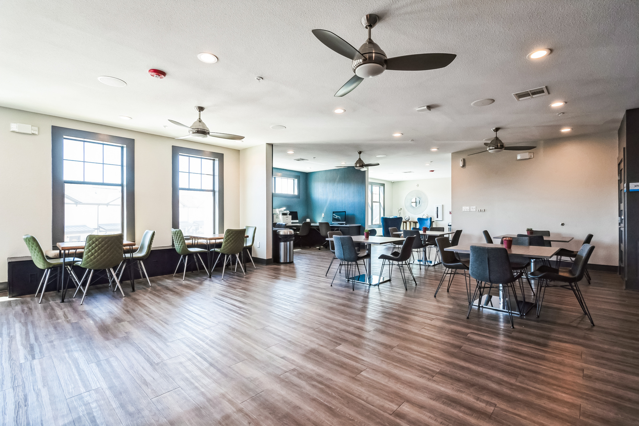Spacious, modern room with wood flooring, multiple tables and chairs, large windows letting in natural light—perfect for TAMU off campus housing—with ceiling fans and a dark accent wall featuring a computer workstation in the corner.