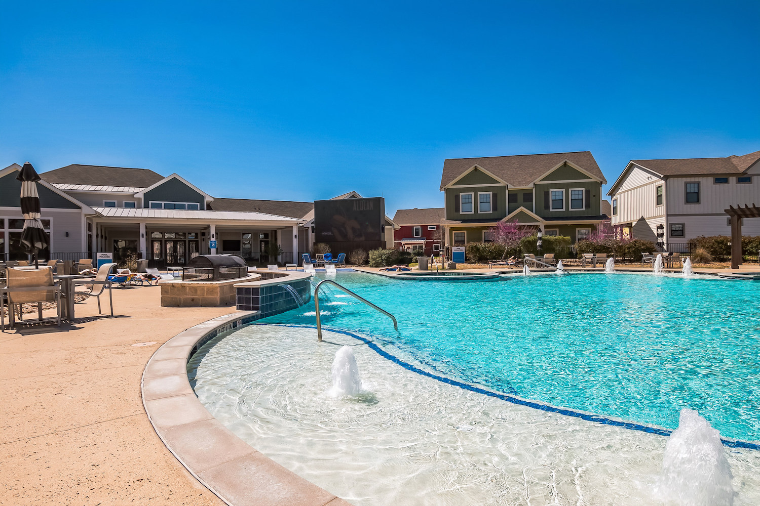 Outdoor swimming pool with fountains, surrounded by lounge chairs and umbrellas, set in apartments in College Station’s residential community with colorful two-story houses and a clubhouse under a clear blue sky.