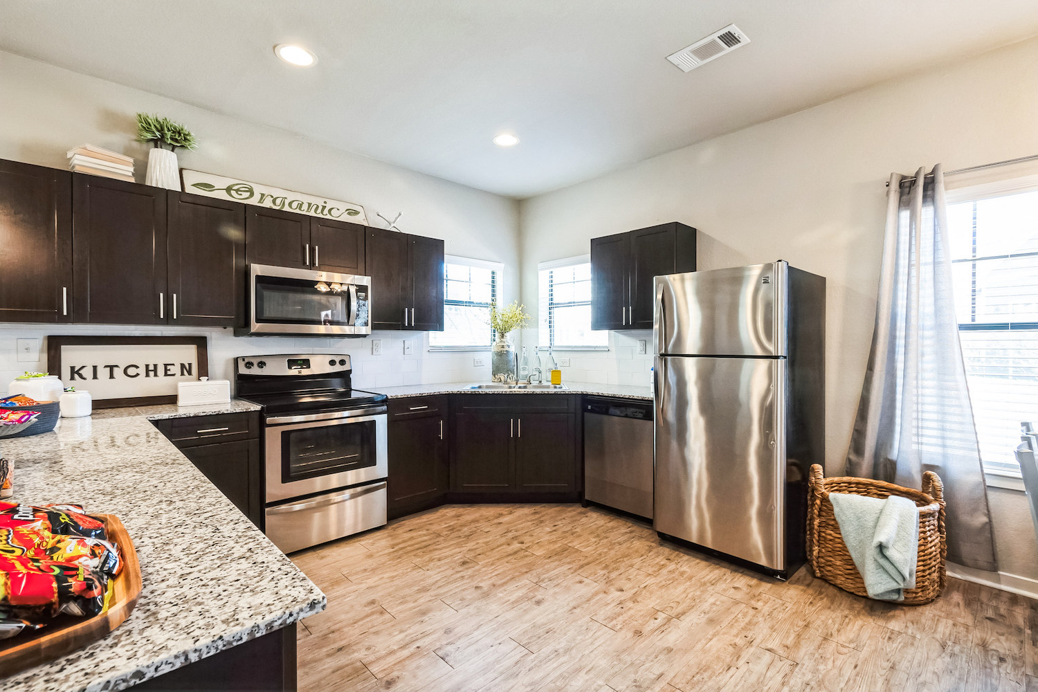 Modern kitchen with dark cabinets, stainless steel appliances, and granite countertops—perfect for Texas A&M freshman housing. Wood-look flooring, natural light, and cheerful decor create a welcoming space; snacks await on the counter by a woven basket of towels.