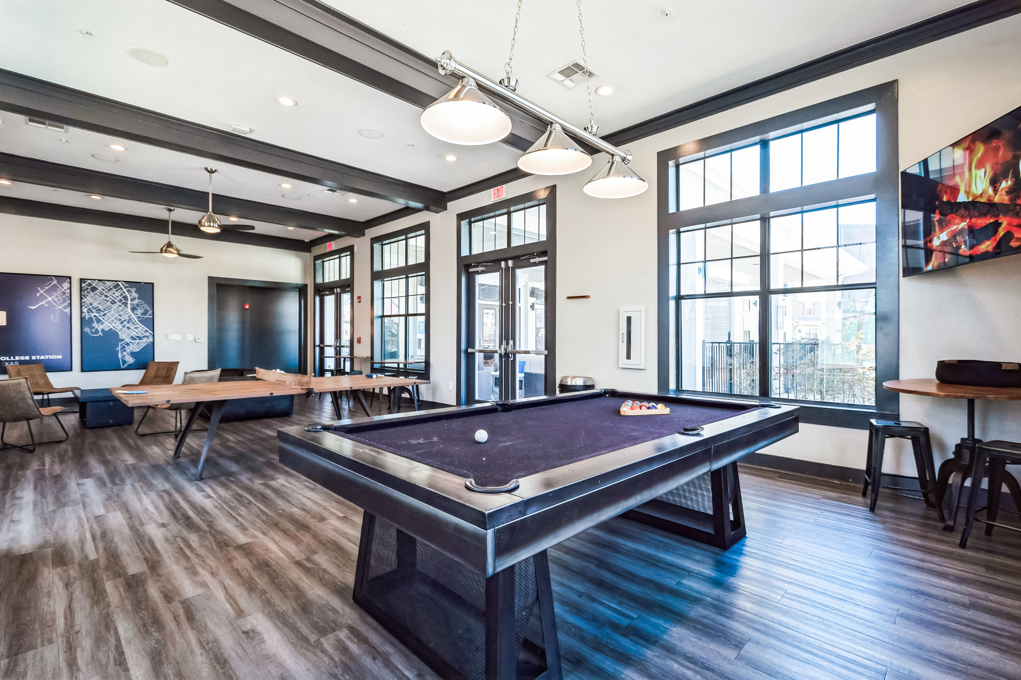 Modern game room in TAMU off campus housing with a black pool table, wooden shuffleboard, lounge chairs, large windows, wood flooring, overhead lights, and a wall-mounted TV displaying a cozy fireplace scene.