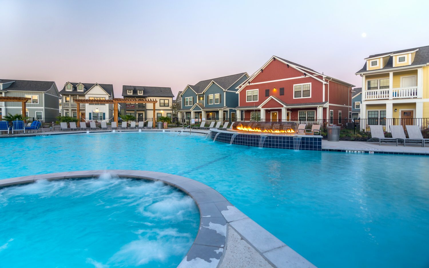 A large outdoor swimming pool with a connected hot tub, surrounded by lounge chairs—ideal for tamu off campus housing. Behind the pool are colorful, multi-story houses and a fire feature, all under a clear evening sky.