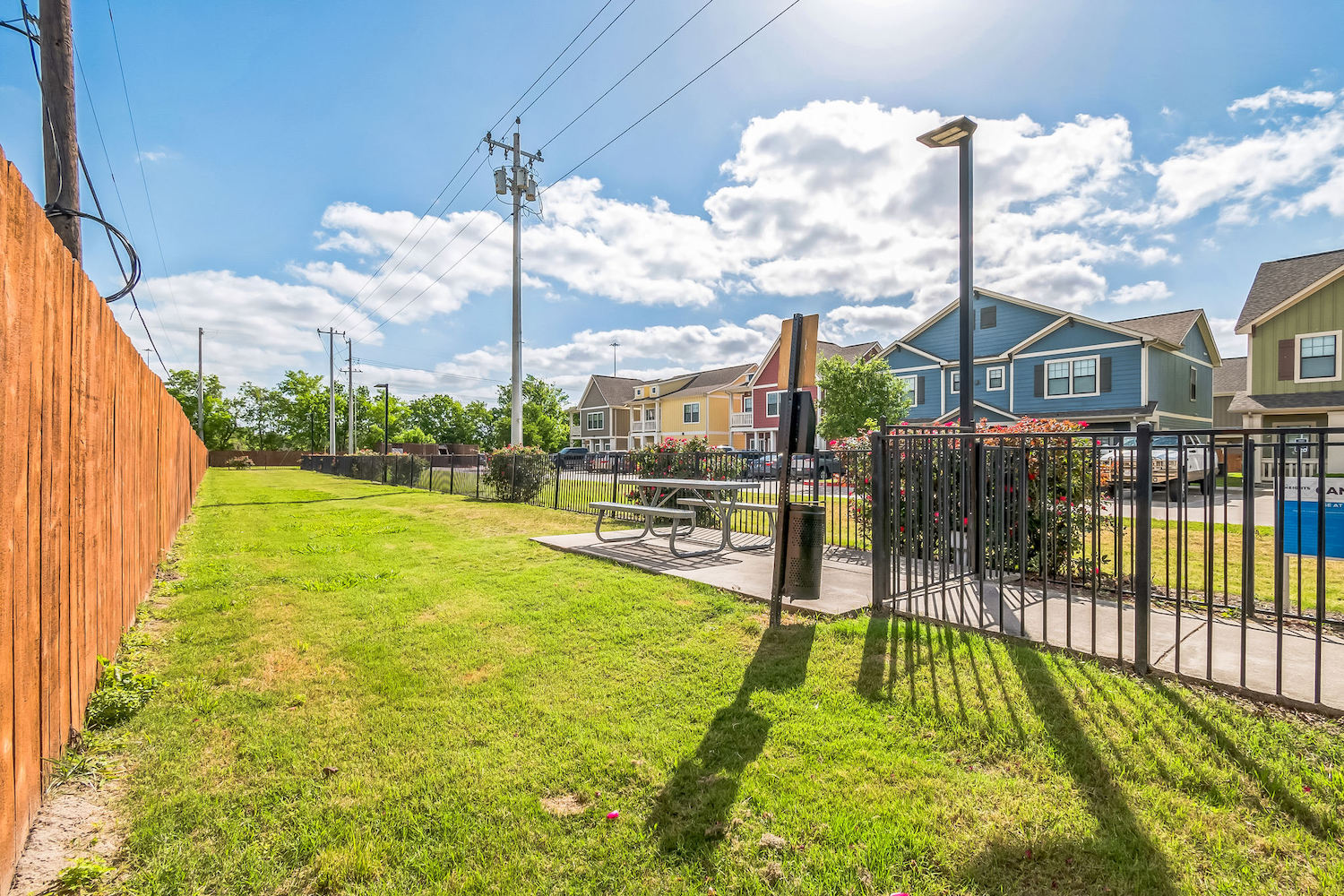 A sunny outdoor area with green grass, a wooden fence, picnic table, grill, and black metal fence—perfect for TAMU off campus housing. Colorful houses and power lines stand out in the background under a blue sky with clouds.