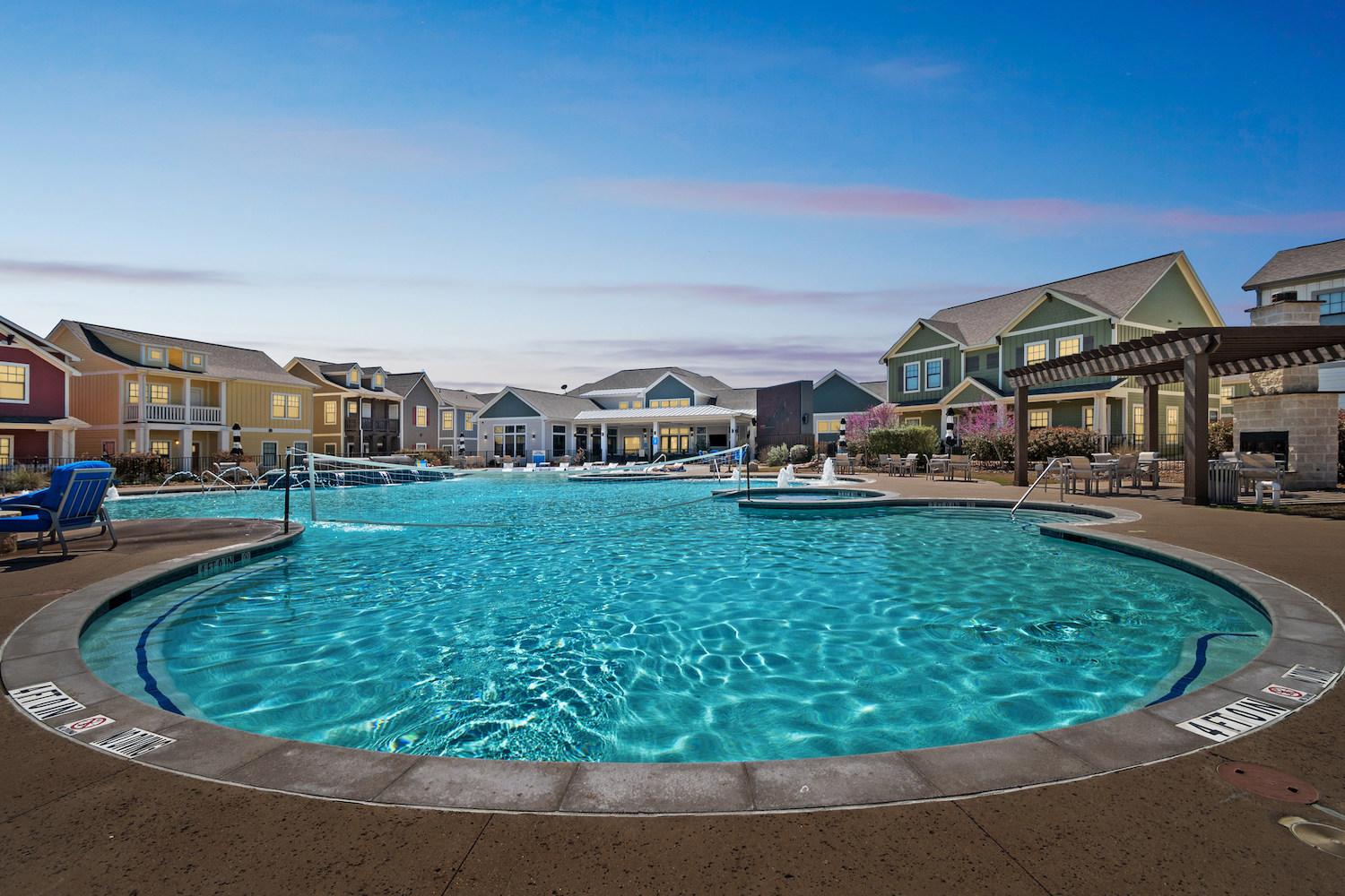Large, circular outdoor swimming pool surrounded by lounge chairs and colorful houses under a clear blue sky at sunset; perfect for Tamu off campus housing, with a pergola and smaller pools visible in the background.