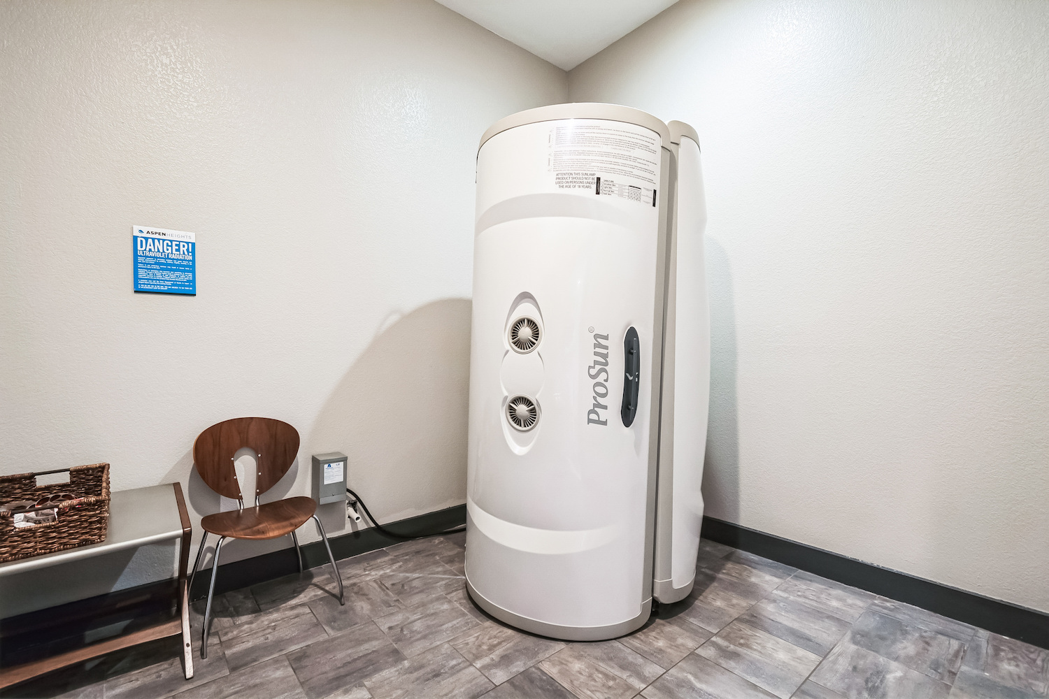 A vertical tanning booth labeled “ProSun” stands in the corner of a small room at Tamu off campus housing, featuring gray walls, a tiled floor, a wooden chair, a basket on a table, and a safety warning sign on the wall.