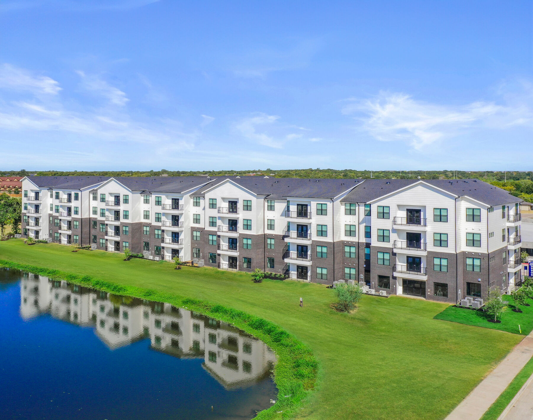 Harbor at the Barracks is a modern four-story apartment building with balconies that overlooks a pond and a large grassy area, surrounded by trees and a blue sky.