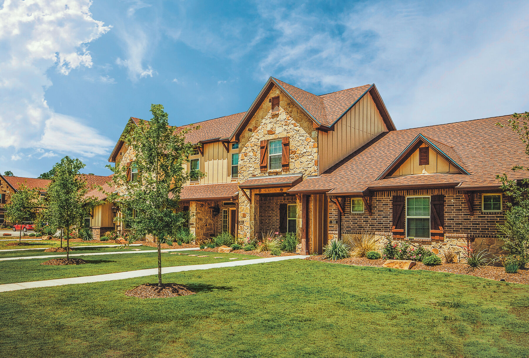 A large, two-story house with brown shingles, stone and brick exterior, green shutters, and a well-maintained lawn with young trees under a bright, blue sky.