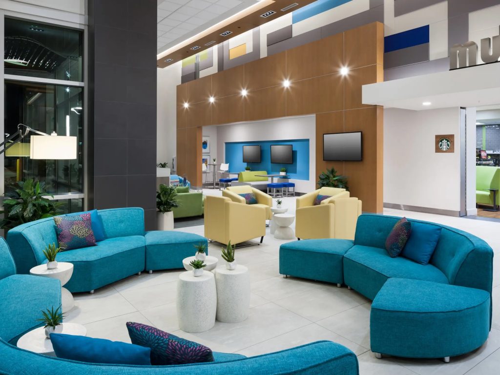 Modern lounge area with curved teal sofas, light green chairs, small white tables with plants, and wall-mounted TVs. The space features bright lighting, colorful wall panels, and a Starbucks counter in the background.