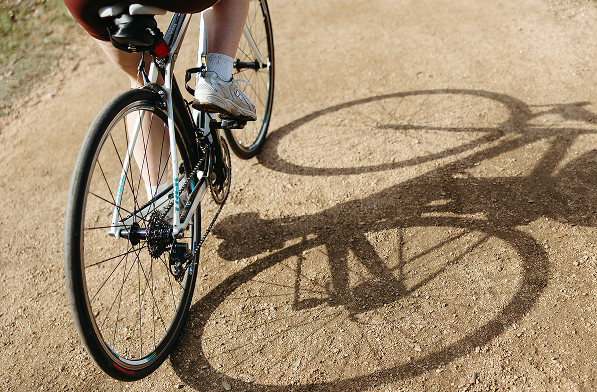 Close-up view of a person riding a bicycle on a dirt path, with their legs and part of the bike visible. The sun casts a sharp shadow of the bike and rider on the ground.