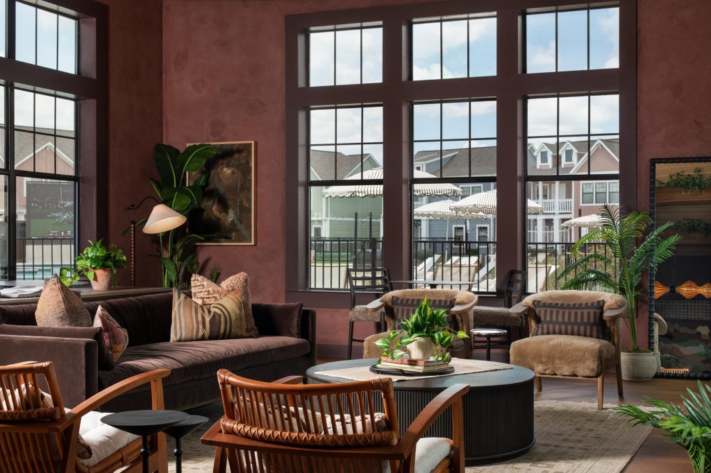A stylish living room with large windows, brown walls, modern furniture, houseplants, and a view of homes and a patio outside—perfect for Texas A&M off campus housing. Light fills the space, creating a cozy, inviting atmosphere ideal for students seeking comfort and convenience.