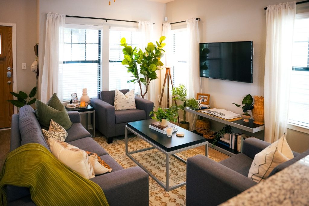 A cozy, sunlit living room with gray sofas from stylish furniture packages The Heights, a large plant, a wall-mounted TV, and a coffee table adorned with candles and greenery. Light curtains and a patterned rug complete the inviting, modern space.