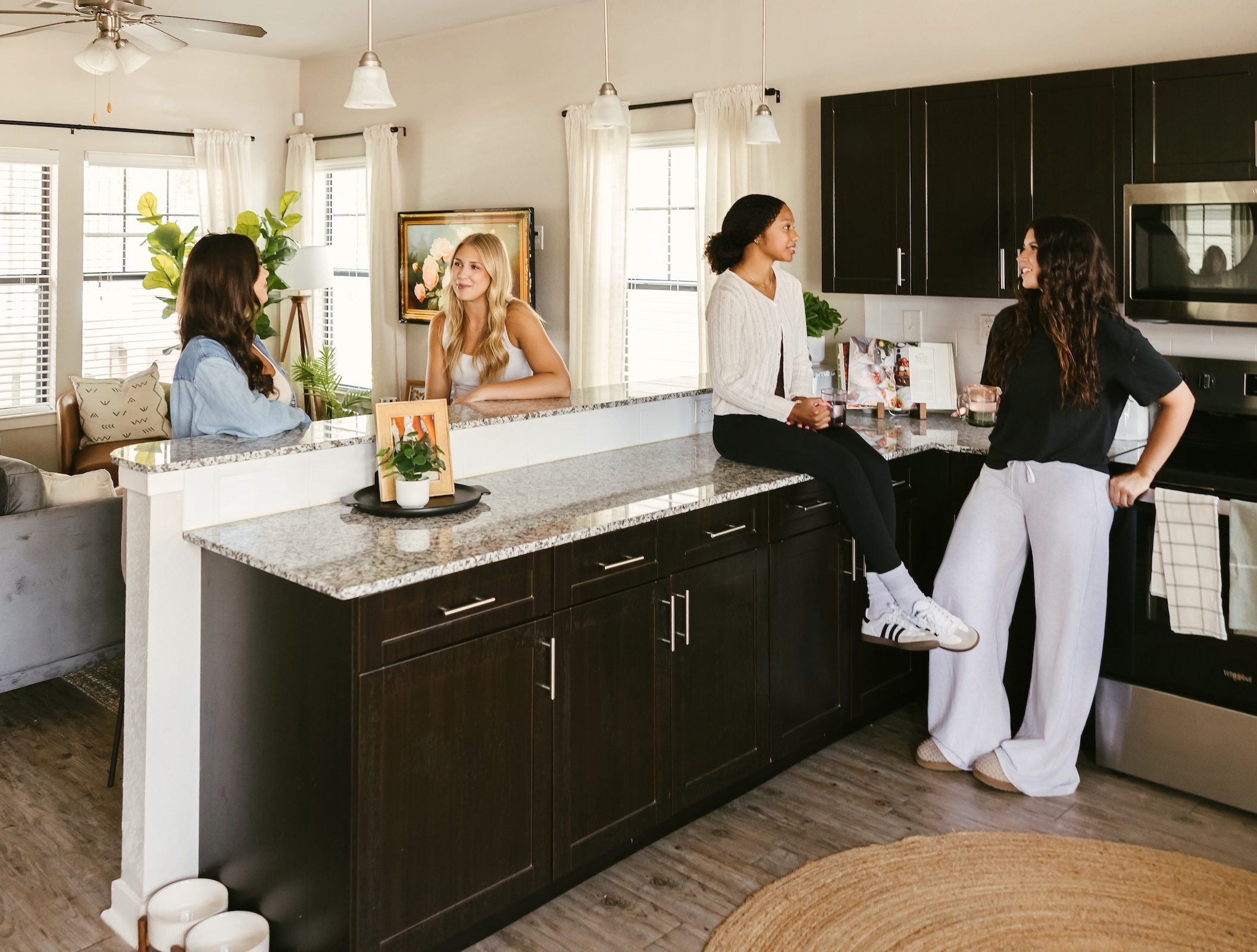 Four women talk and smile in a bright, modern kitchen with dark cabinets and granite countertops. One sits on the counter, two stand, and one leans in from the living room, creating a warm, social atmosphere.