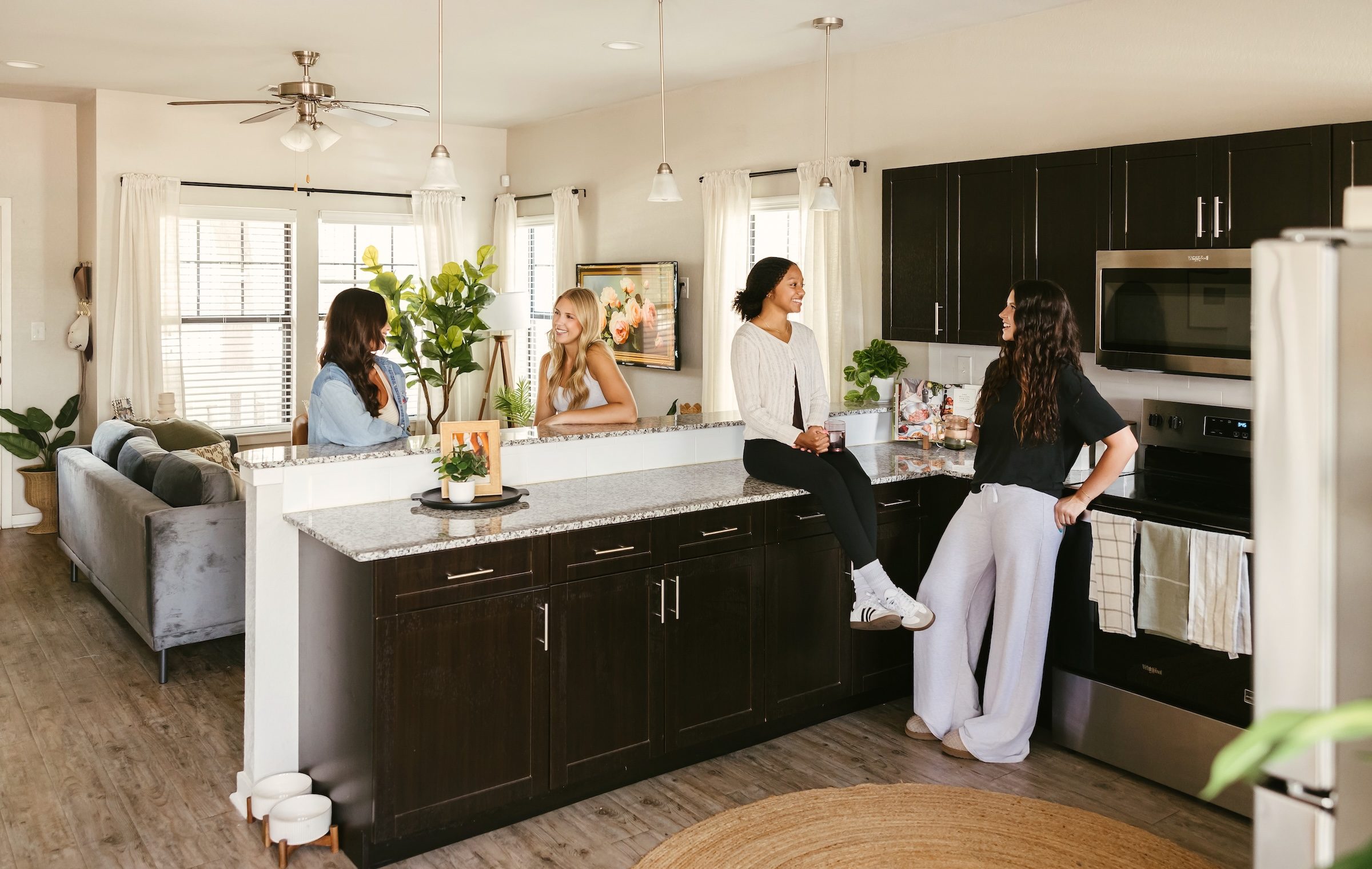 Four women are talking and smiling in a modern kitchen. Two sit on the counter while the other two stand nearby. The space is bright, with dark cabinets and light countertops. A living room with a sofa is visible in the background.