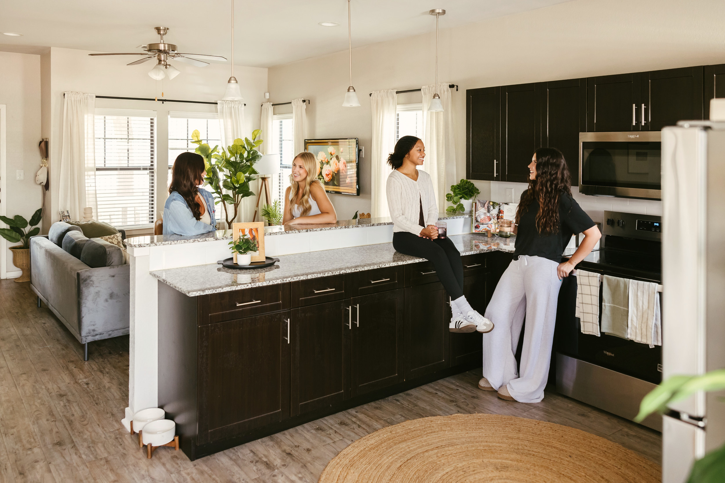 Four women chat and smile in a modern kitchen with dark cabinets and stainless steel appliances, giving the space a polished look reminiscent of upscale Texas A&M freshman housing; a cozy living room is visible in the background.