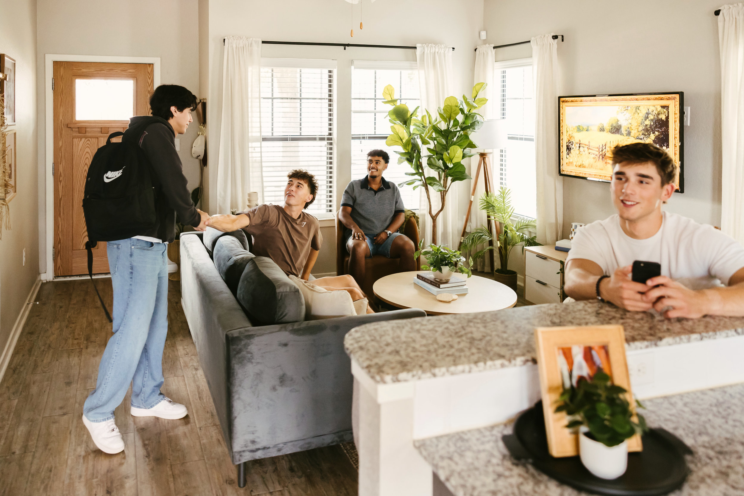 A person with a backpack shakes hands with someone on a couch—a welcoming scene reminiscent of Texas A&M freshman housing. Two other people sit nearby, one holding a phone, in a bright living room with plants and a TV on the wall.