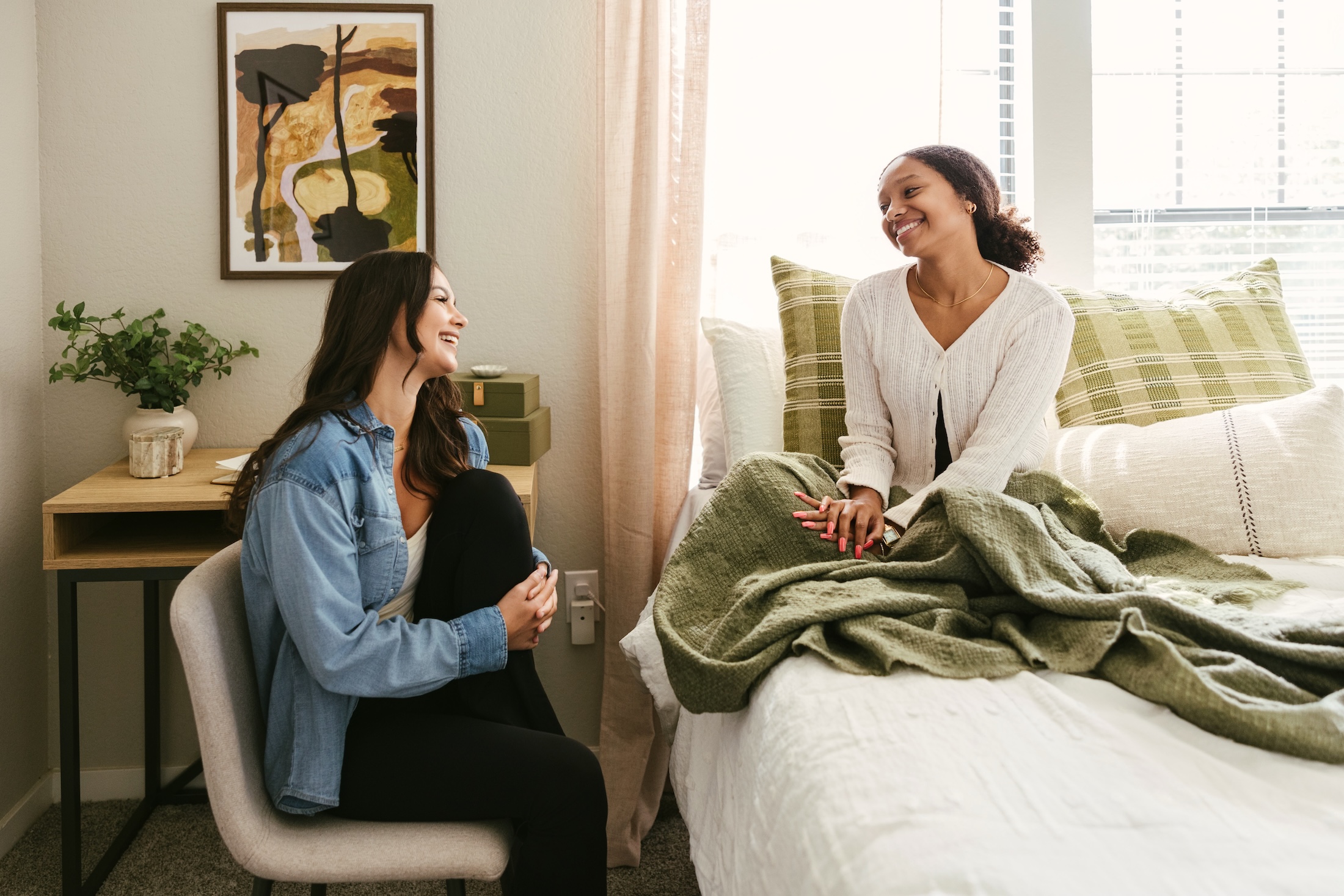 Two women smile and talk together in a cozy bedroom. One sits on a chair, the other sits on a bed with a green blanket. Sunlight shines through a window with blinds, and theres a plant and art on the wall.