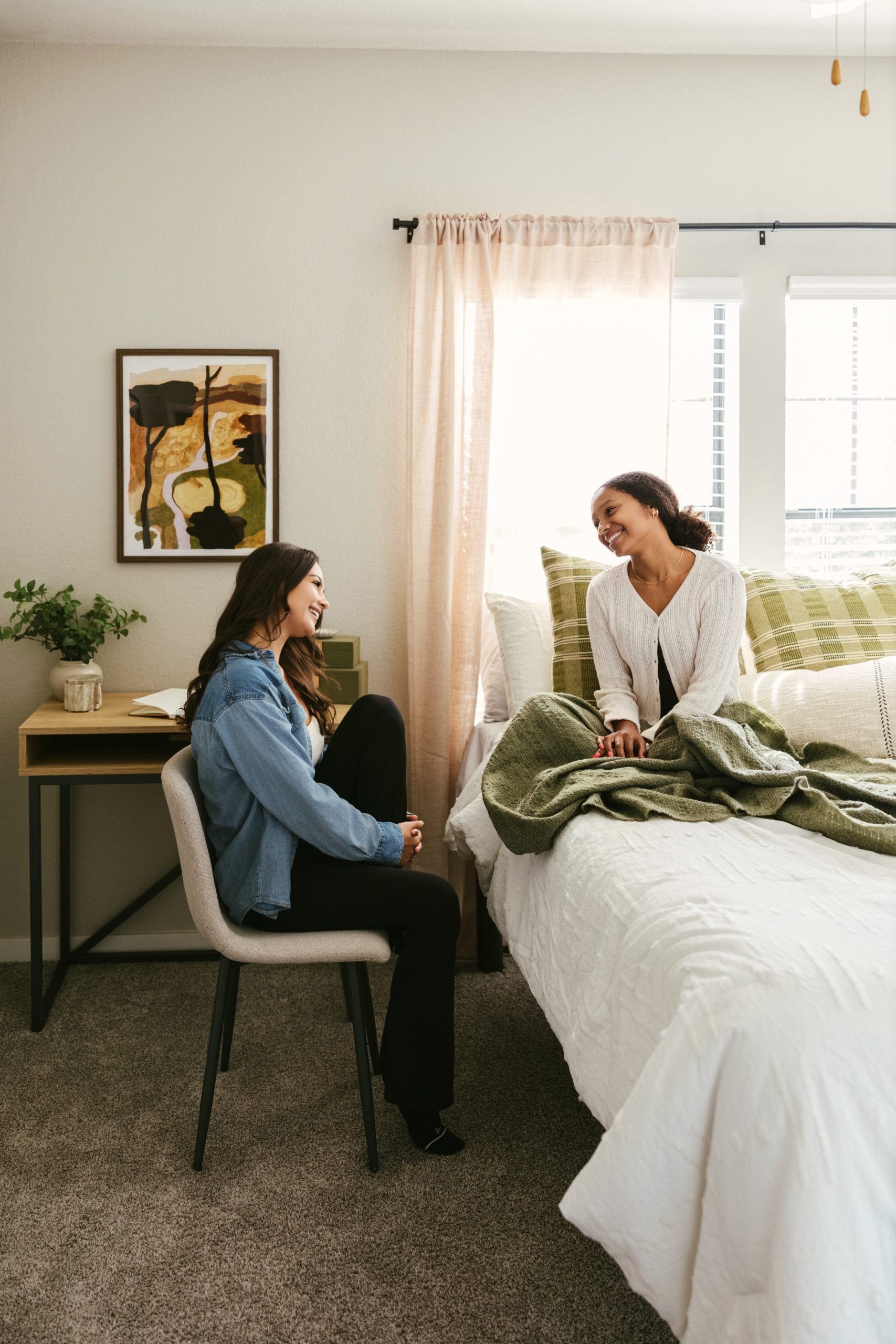 Two women smiling and talking in a cozy bedroom; one sits on a chair by a desk, the other sits on a bed with a green blanket. Sunlight filters through sheer curtains behind them, highlighting the thoughtful décor inspired by furniture packages at The Heights.