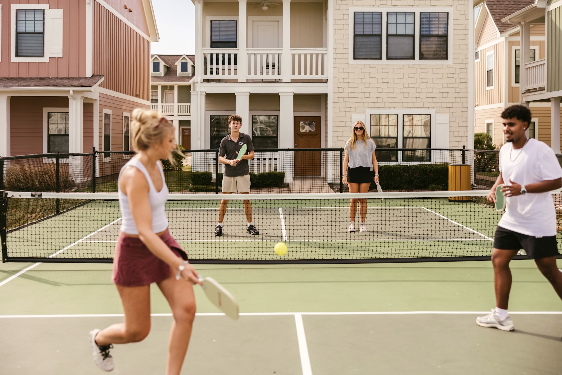 Four young adults play pickleball on an outdoor court in front of suburban houses. Two are near the net, ready to hit the ball, while the other two stand on the opposite side, watching.