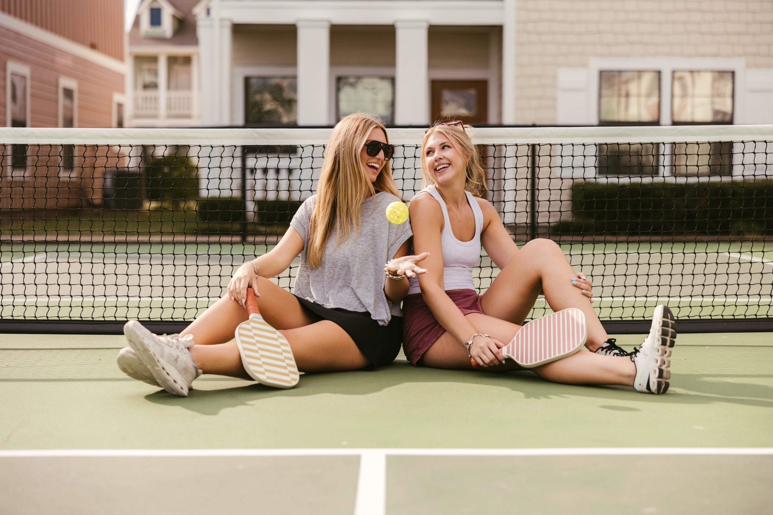 Two women sit on a tennis court, smiling and holding pickleball paddles. One wears sunglasses and tosses a ball in the air. The net and buildings are visible in the background.
