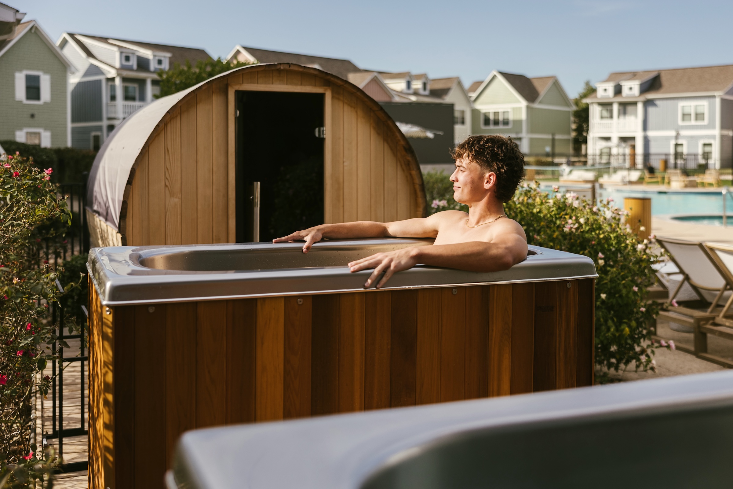 A person relaxes in an outdoor wooden cold plunge near a sauna, with residential houses and a pool visible in the background on a sunny day.