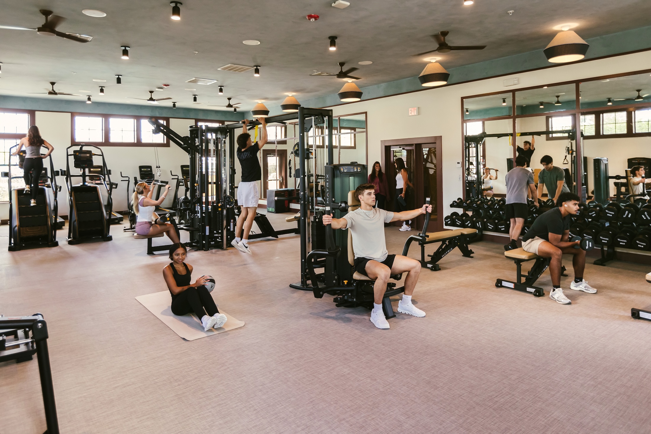 A group of people exercise in a gym, using various equipment like treadmills, weights, and machines. Some are lifting weights, others are using cardio machines, and one person is stretching on a mat.