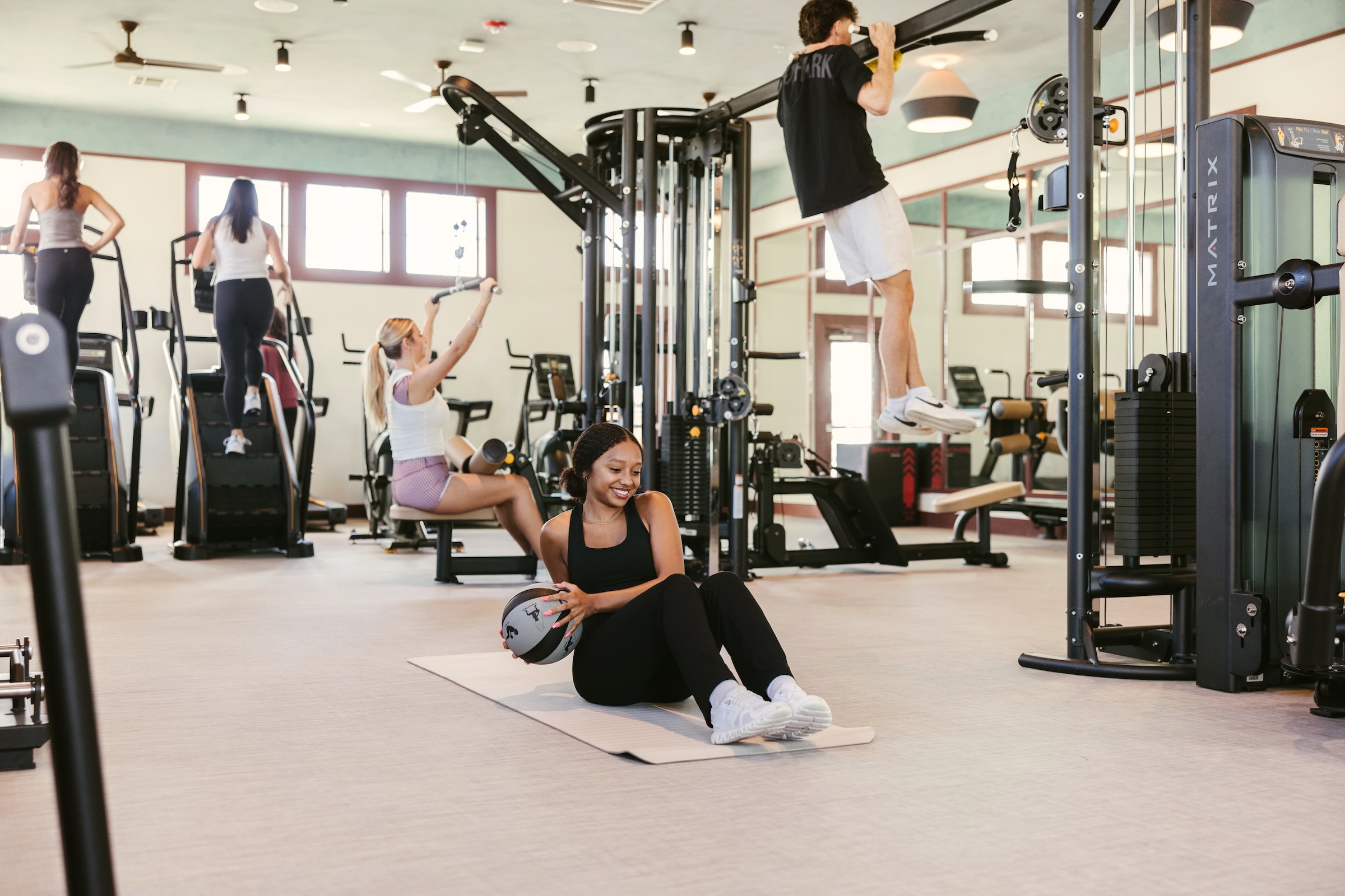 Four people exercise in a gym with various equipment. One person does sit-ups with a medicine ball on a mat, one uses resistance bands, one lifts weights, and one does pull-ups. The gym is bright with large windows.