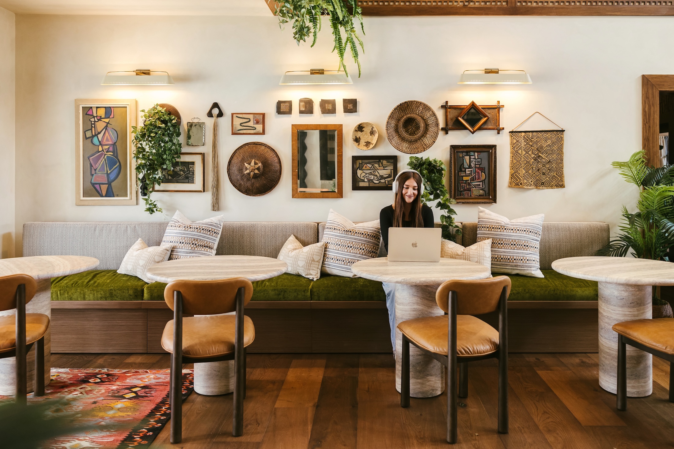 A woman sits with a laptop at a round table in a cozy, stylish cafe with wall art, plants, and cushioned bench seating.