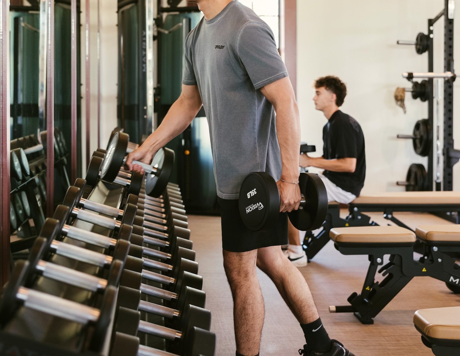 A man in a gray shirt lifts a dumbbell at a gym with mirrors and large windows. Another person sits on a bench in the background, also exercising.