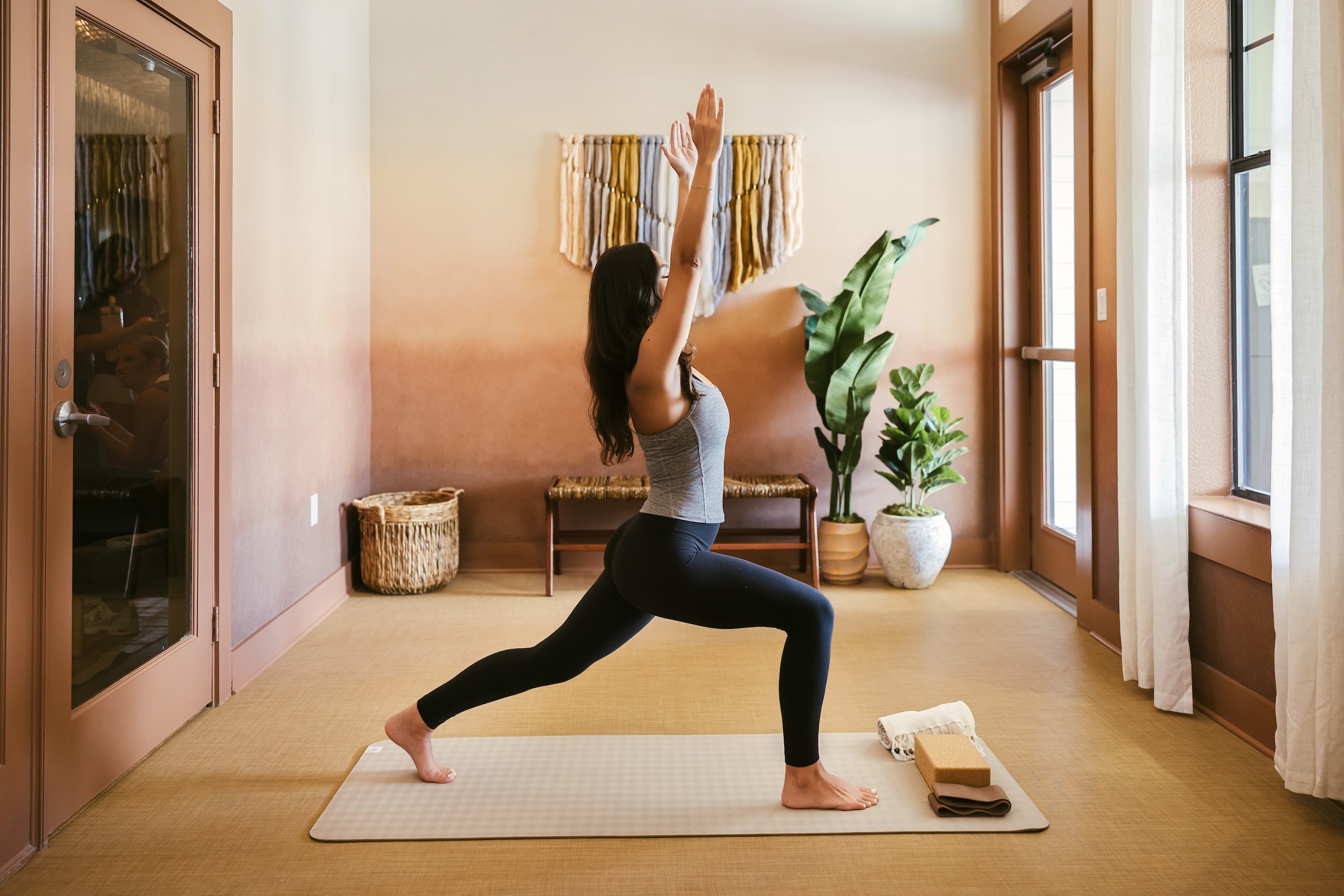 A woman practices yoga in a bright room, performing a high lunge pose on a mat. She faces a window, arms raised, with plants and decorative items in the background.