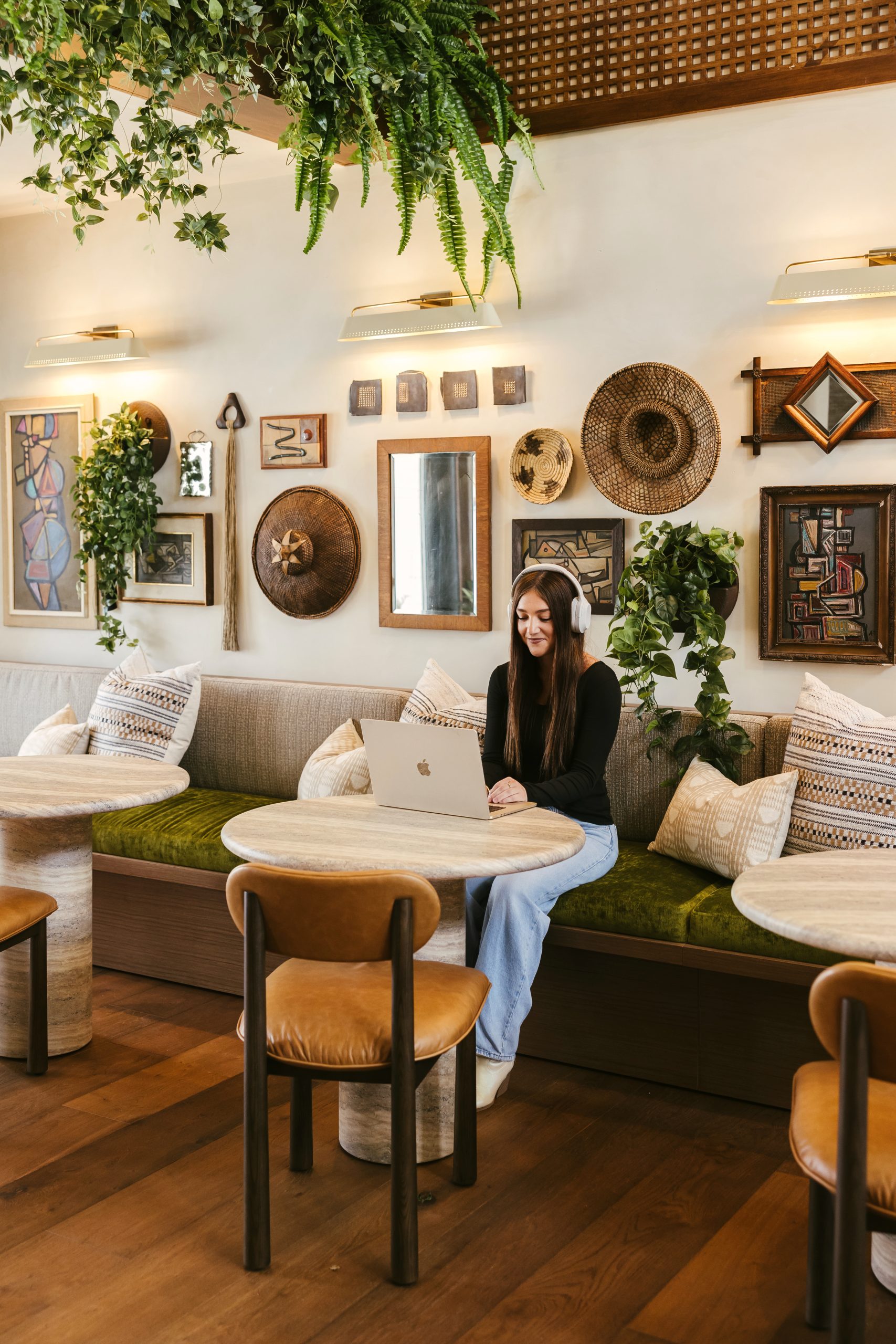 A woman with long hair sits at a round table in a cozy cafe, working on a laptop—perhaps searching for apartments in College Station. The wall behind her is decorated with plants, woven baskets, and framed art. The space has warm lighting and wooden furniture.