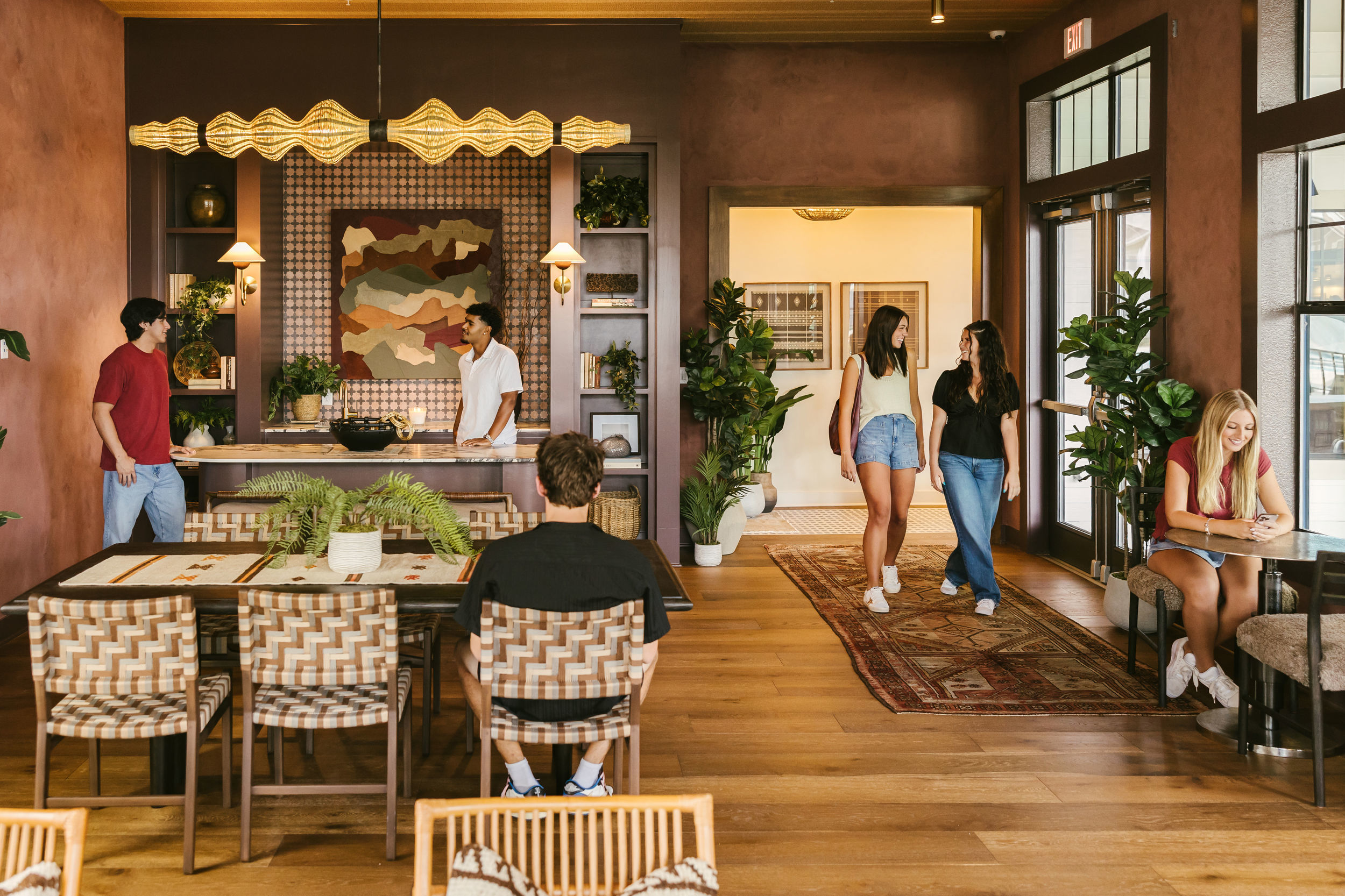 Five young adults are in a stylish, modern lounge at The Heights College Park, with wooden floors, plants, and vibrant wall art. Some are chatting, one is seated at a table, and another writes in a notebook by the window. The atmosphere is casual and welcoming.