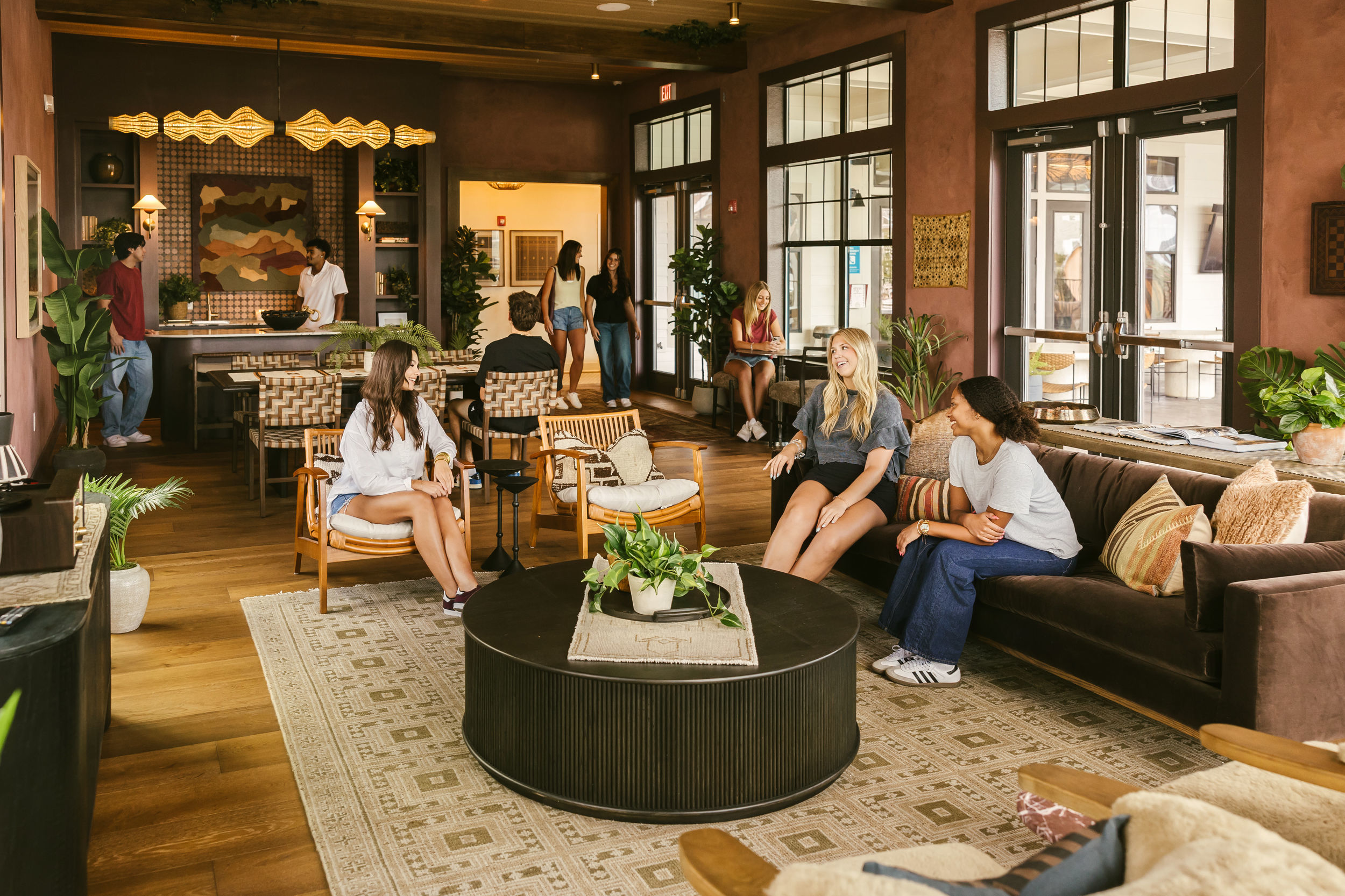 People relax and chat in a stylish, modern lounge at The Heights College Park, with wooden floors, large windows, plants, and comfortable seating. Some are sitting on couches and chairs, while others stand by a counter in the background.