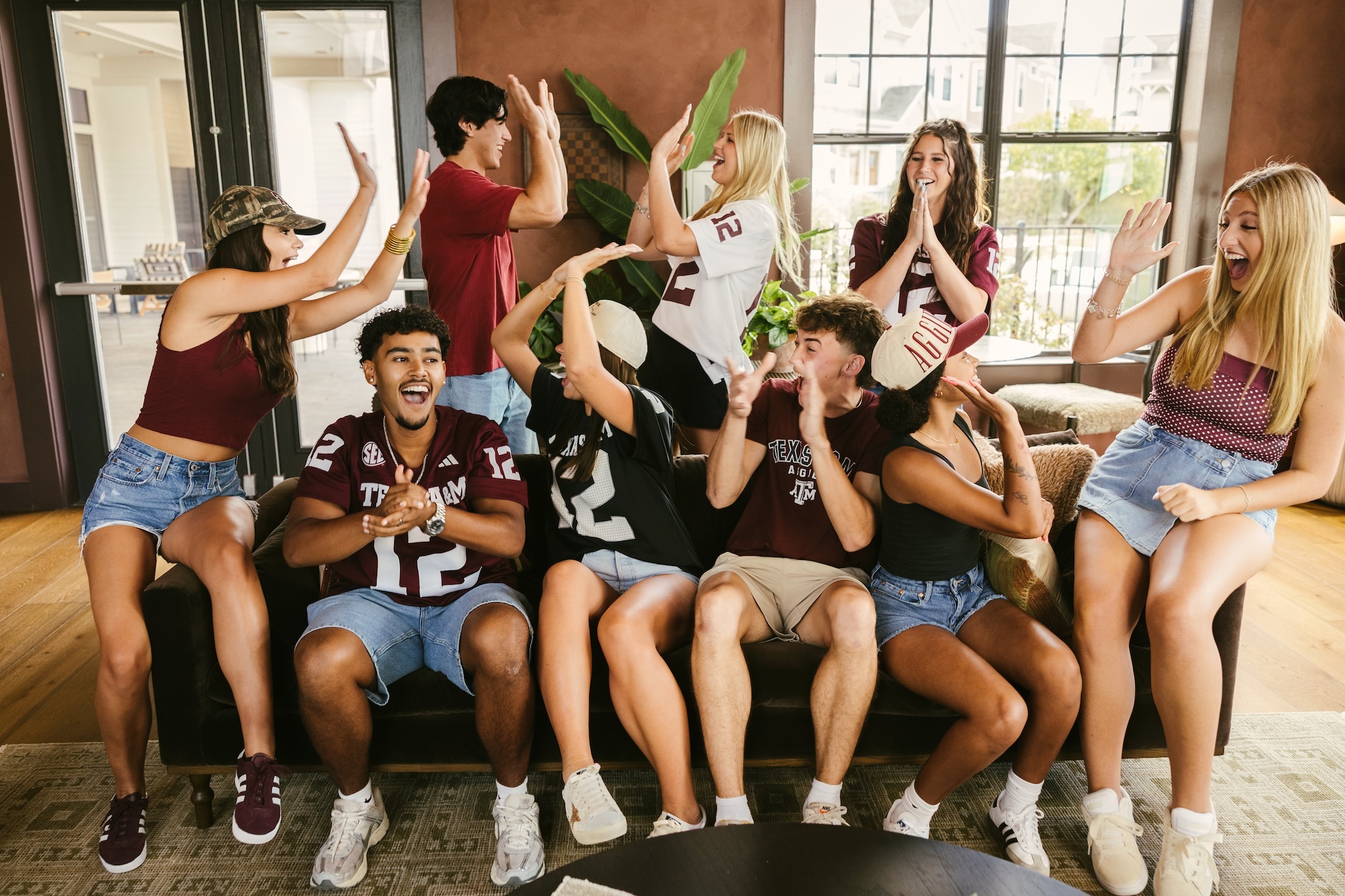 A group of young adults wearing maroon and denim cheer and high-five in a living room, celebrating together on and around a sofa near large windows.