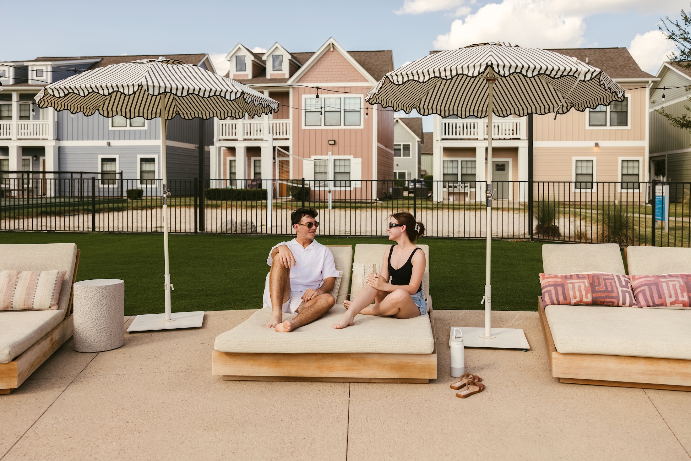 A man and a woman sit and talk on lounge chairs by a pool, under striped umbrellas. Colorful houses and a black fence are in the background. The weather is sunny with a few clouds.