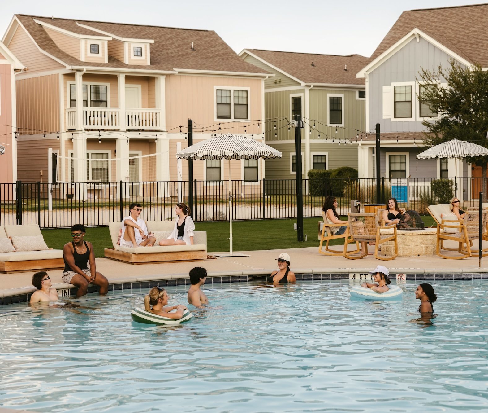 People relax at a pool in a suburban neighborhood. Some swim or float in the water, while others sit on poolside chairs under umbrellas. Colorful houses and a lawn are visible in the background.