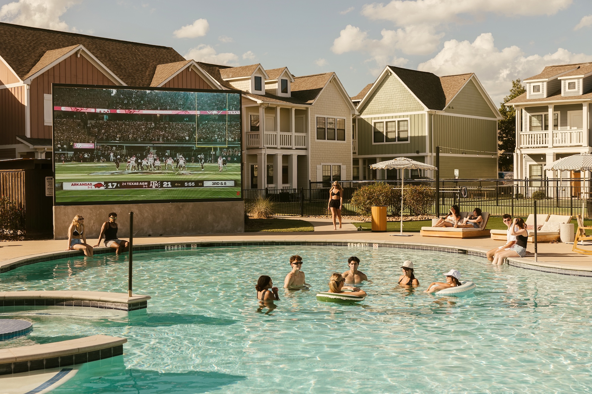 People relax in a community pool while a large outdoor screen shows a football game. Some sit on pool floats, others lounge by the poolside, with houses and a sunny sky in the background.
