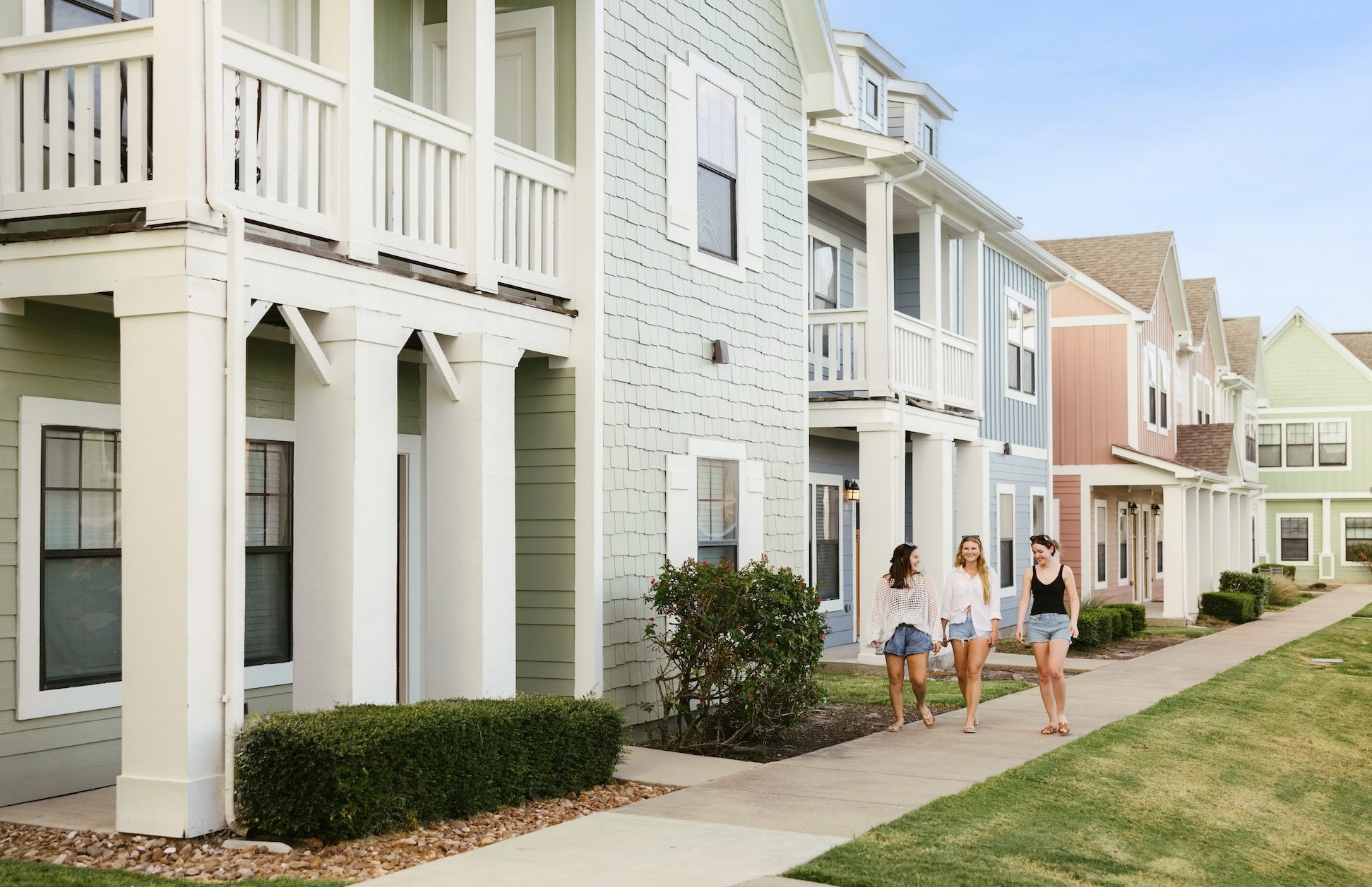 Three young women walk and talk on a sidewalk in front of pastel-colored townhouses with porches on a sunny day.