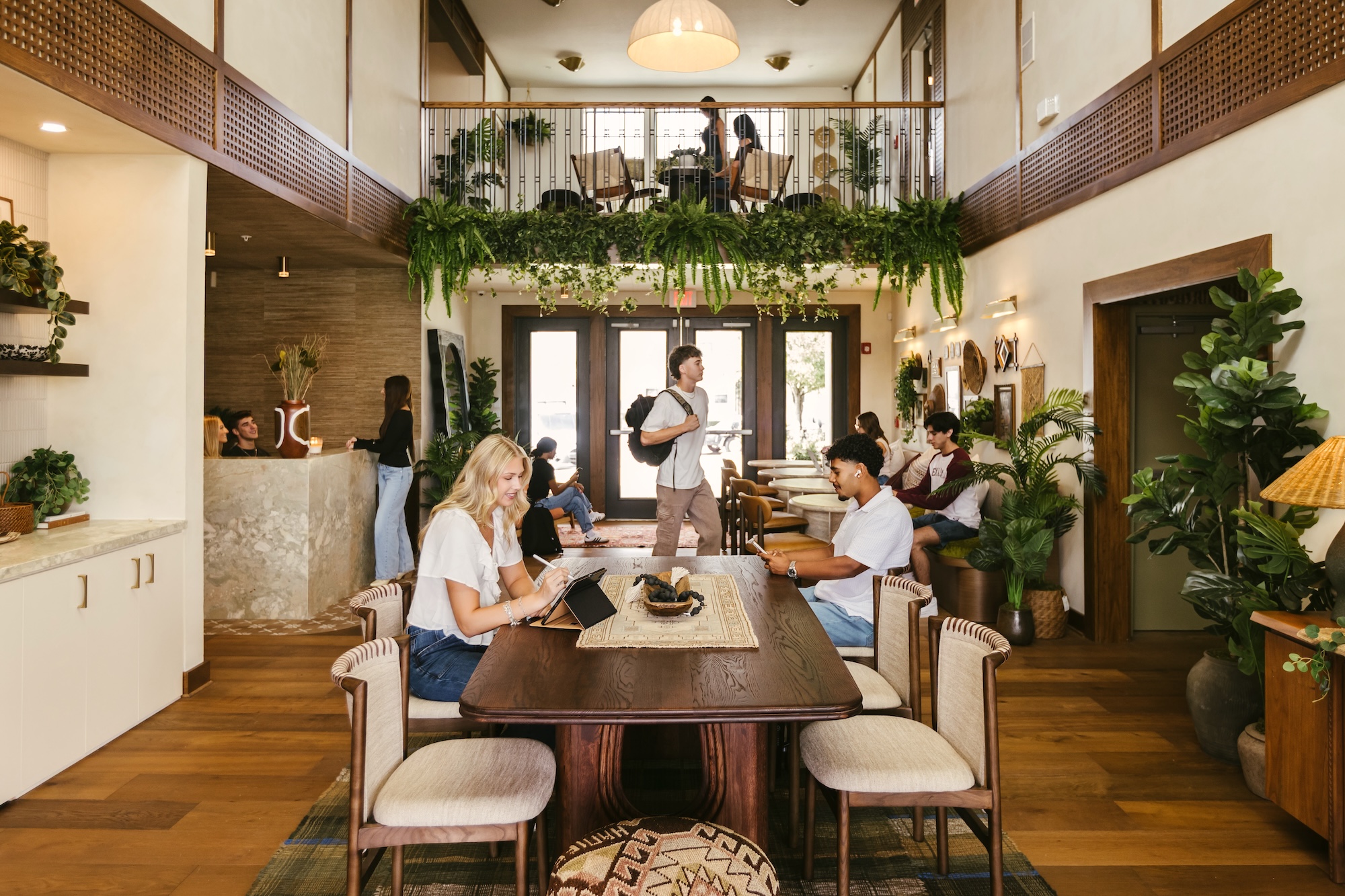 Modern coworking space with people working, talking, and using laptops at tables. Green plants hang from a balcony, and natural light brightens the room with wooden floors and cozy decor.